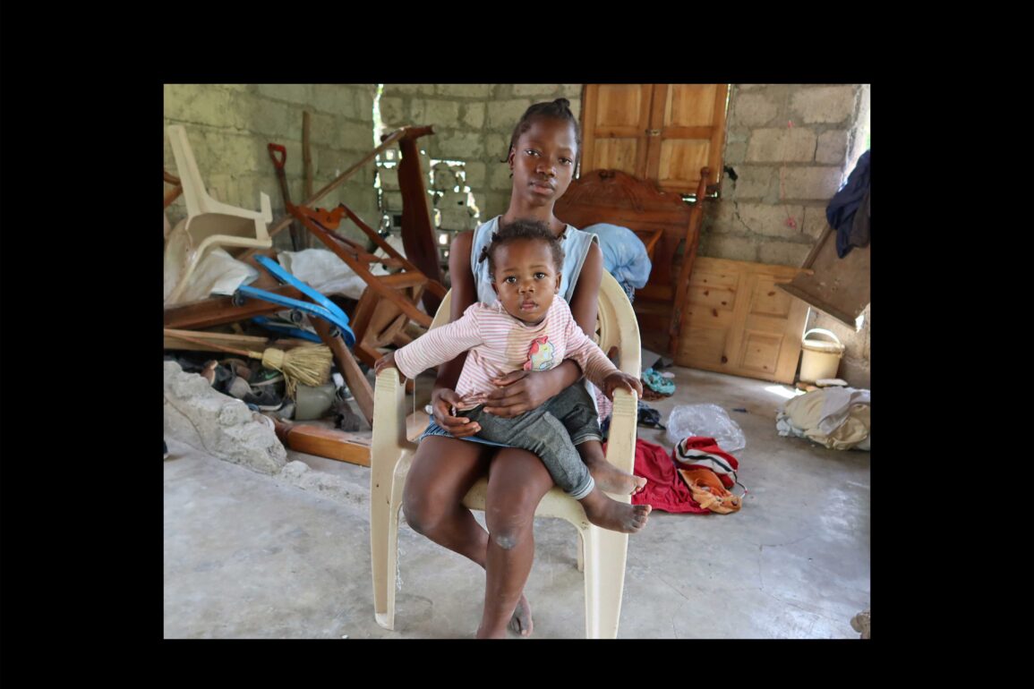 Ein junges Mädchen sitzt in einem zerstörten Haus auf einem Stuhl und hat ein Kind auf ihrem Schoss.